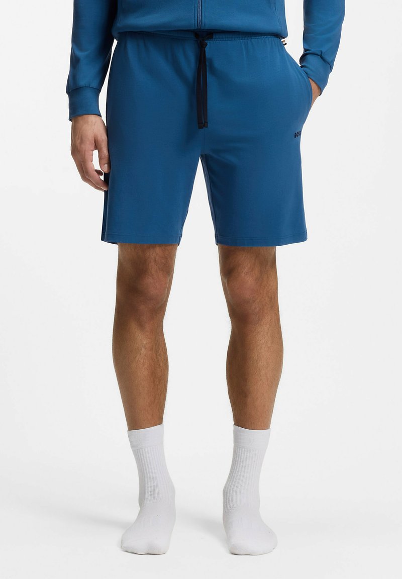 Homme portant un short bleu à cordon de serrage et une veste bleue assortie, avec des chaussettes blanches montantes, debout devant un fond blanc uni.