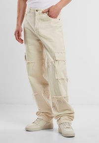 Pantaloni cargo bej într-o croială lejeră, cu accente desfăcute, multiple buzunare aplicate și un material texturat. Se asortează cu adidași de culoare deschisă.