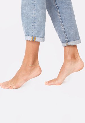 Gold ankle bracelet with a small heart charm, worn on bare foot. Foot is partially posed, paired with rolled-up light blue denim jeans.