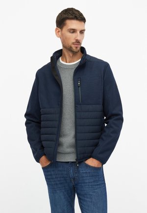 Hombre con chaqueta acolchada azul marino sobre suéter gris y vaqueros azules, de pie con las manos en los bolsillos, mirando hacia abajo y hacia un lado.
