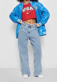 Modrá zateplená bomber bunda s bílými pruhy, červený krátký svetr s nápisem "USA" a světle modré široké džíny s vyšívaným logem.