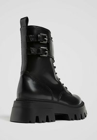 Črni usnjeni gležnjar z dvema nastavljivima zaponkama, okrogel prst, debela platforma podplat in teksturirana površina. Ima vezalko spredaj.