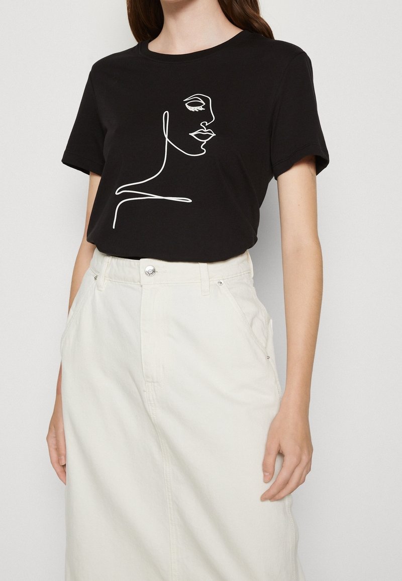 Femme portant un T-shirt noir avec un dessin en ligne blanche continue d’un visage féminin, associé à une jupe blanche taille haute.