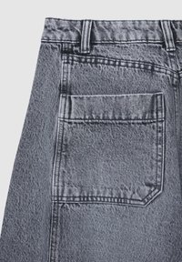 Detail der hinteren Tasche von ausgebleichten grauen Denim-Jeans mit sichtbaren Nähten und Gürtelschlaufen auf einem hellen Hintergrund.