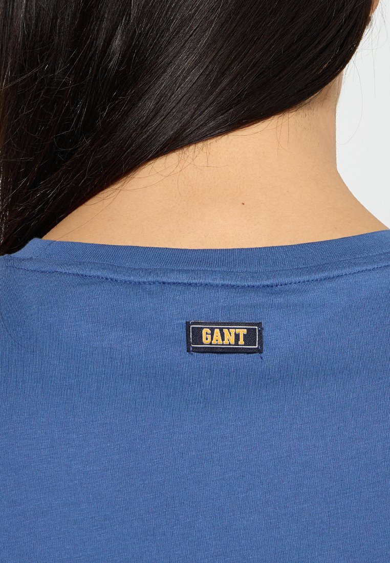 Blaues T-Shirt aus glattem Stoff, mit rundem Halsausschnitt und einem kleinen schwarzen und gelben "GANT"-Logosticker auf der Rückseite.