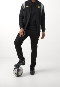 Svart Puma träningsdräkt med gula detaljer och logotyp, svarta fotbollsskor och grå detaljer, ståendes på en svart och vit fotboll.