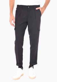 Pantaloni cargo neri, sartoriali, con pieghe, tasche laterali e orli a giro, realizzati in un tessuto liscio. Abbinati a sneakers bianche.