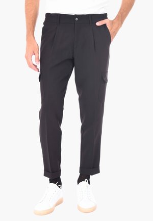 Pantaloni cargo neri, sartoriali, con pieghe, tasche laterali e orli a giro, realizzati in un tessuto liscio. Abbinati a sneakers bianche.