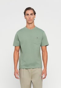 Man in een saliegroen T-shirt met korte mouwen en een logopocket op de borst, met beige broek, staand tegen een effen witte achtergrond.