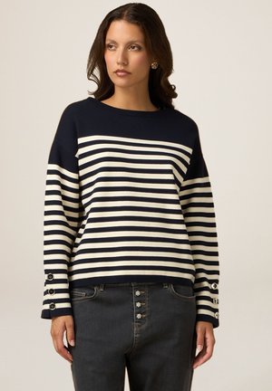 Maglione a righe navy e crema con scollatura rotonda, spalle scese e dettagli in bottone sulle maniche. Abbinato a jeans scuri.