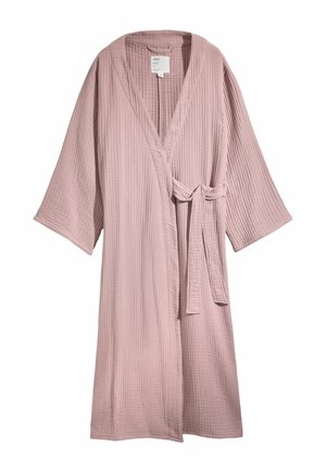 Nő, aki egy texturált, poros rózsaszín átvetős köntöst visel szürke felső fölött, szabadon lógó hajjal, amely részben takarja az arcát, világos fal előtt.