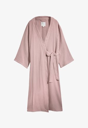 Vrouw draagt een gestructureerde stoffige roze wikkelbadjas over een grijs topje, met los haar dat gedeeltelijk haar gezicht bedekt, tegen een lichte muur.