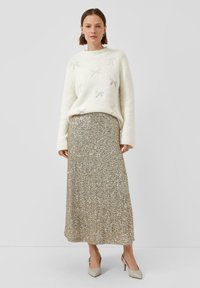Vrouw in een witte trui met strikpatronen, een gouden pailletten midi-rok en beige puntige hakken, staand tegen een effen achtergrond.