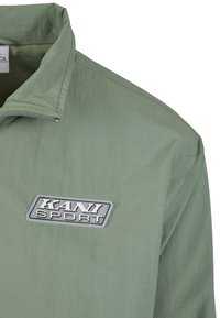 Šviesiai žalia striukė su užtrauktuku ant apykaklės, pasižyminti tekstūruota medžiaga ir ryškiu pilku „KANI SPORT“ logotipu krūtinėje.