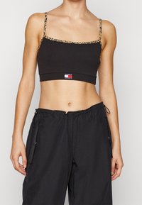 Donna che indossa un top nero cropped con rifiniture in stampa leopardo e pantaloni neri larghi con tasche laterali, su uno sfondo neutro.