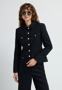 Czarna marynarka o dopasowanym kroju z kołnierzykiem, złotymi guzikami i wieloma kieszeniami z przodu, w zestawie z czarnymi spodniami. Gładka faktura, usztywniony fason.