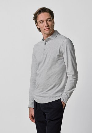 Mężczyzna ubrany w jasnoszarą koszulkę polo z długim rękawem i czarne spodnie, patrzący w lewo, z jedną ręką w kieszeni, na jednolitym tle.