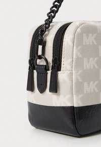 Kanjus maišelis smėlio spalvos su baltu MK logotipu, juodu dugnu, dviem užtrauktukais ir juoda grandinėlės petnešėlėmis. Textūruotos paviršiai ryškiai kontrastuoja.