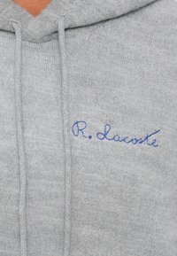 Haut à capuche gris en tissu doux avec une poche avant, arborant une écriture brodée bleue "R. Lacoste" sur le côté gauche.