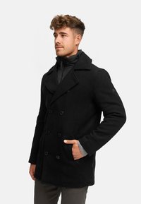 Schwarzer doppelreihiger Peacoat mit großen Knöpfen, Wollstoff, breitem Kragen und frontalen Taschen, über einer schwarzen Daunenjacke getragen.