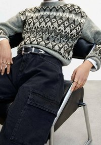 Sweter dzianinowy w kolorze szarym z czarno-białymi wzorami geometrycznymi, połączony z czarnymi spodniami typu cargo i paskiem ozdobionym srebrnymi ćwiekami.