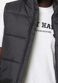 Šedá prošívaná vesta s zipem, skládající se z prošívaných sekcí. Nositelná přes bílé tričko s černým nápisem. Látka vypadá hladká a lehká.
