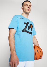 Světle modré tričko Nike s krátkým rukávem, které má velkou černou grafiku s oranžovým designem basketballu. Hladká textura, volný střih.