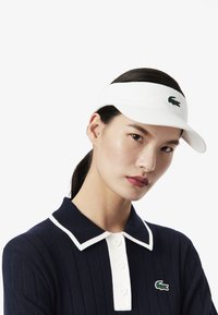 Visière blanche avec une bande solide, arborant un logo Lacoste vert. Le modèle porte un polo bleu marine avec des accents blancs et une texture côtelée.