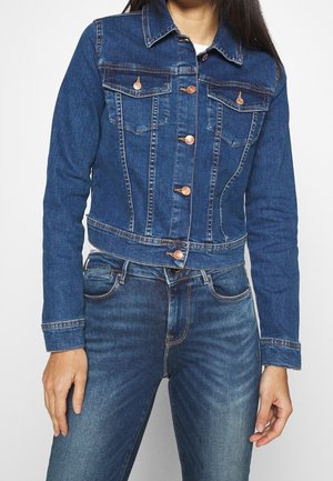 Femme portant une veste en jean bleue ajustée avec des boutons en cuivre et un jean bleu assorti sur un fond blanc uni.