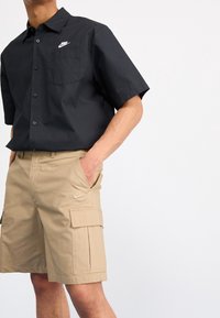 Svart kortärmad skjorta med ficka och Nike-logotyp; beige cargoshorts med cargofickor och Nike-logotyp. Båda i lättviktsmaterial.