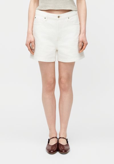 Femme portant un short en jean blanc, des chaussures Mary Jane marron et un haut rayé court, debout devant un fond clair uni.