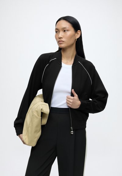 Femme aux longs cheveux noirs portant une veste noire zippée sur un haut blanc, tenant un sac beige, regardant sur le côté sur un fond uni.