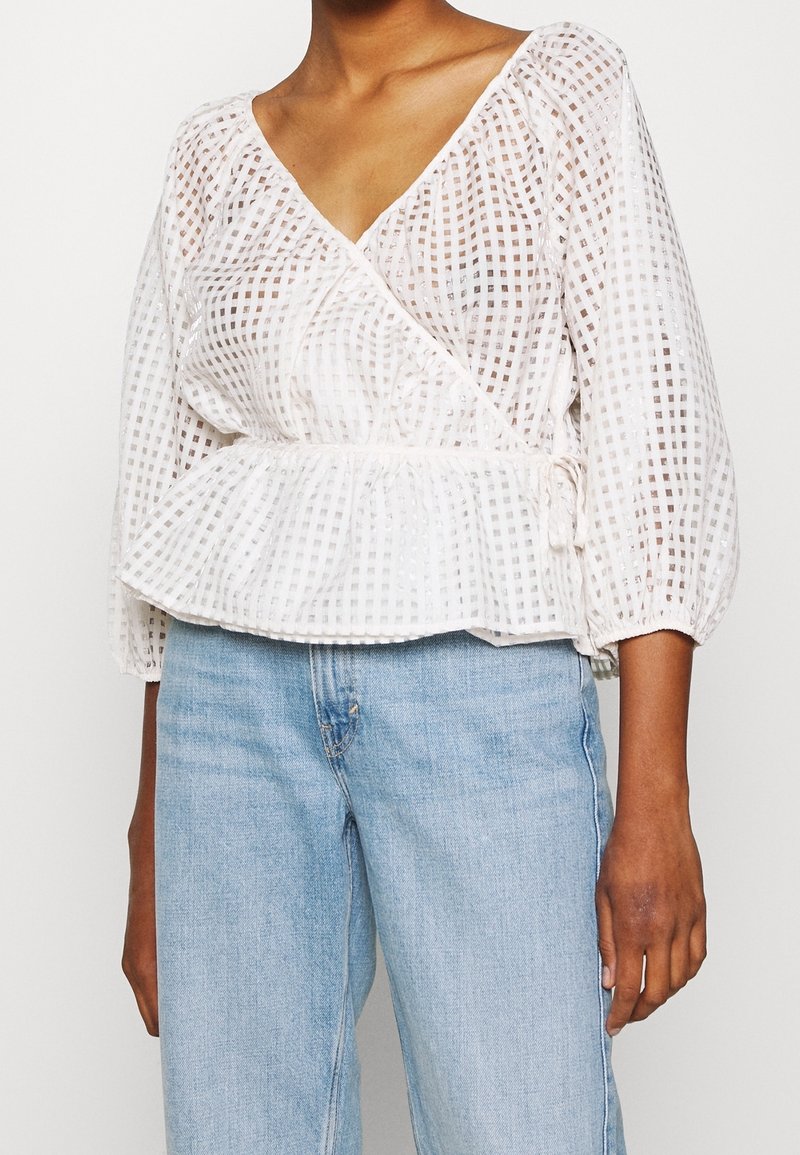 Personne portant un chemisier cache-cœur blanc semi-transparent à carreaux avec des manches bouffantes 3/4 et un jean taille haute bleu clair.