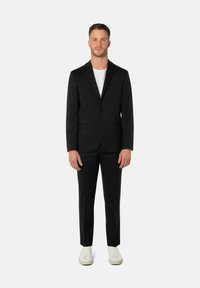 Schwarzer Anzug mit maßgeschneiderter Passform, einreihigem Blazer mit einem Knopf, aufgesetzten Revers und Seitentaschen, kombiniert mit weißem T-Shirt und lässigen weißen Sneakers.