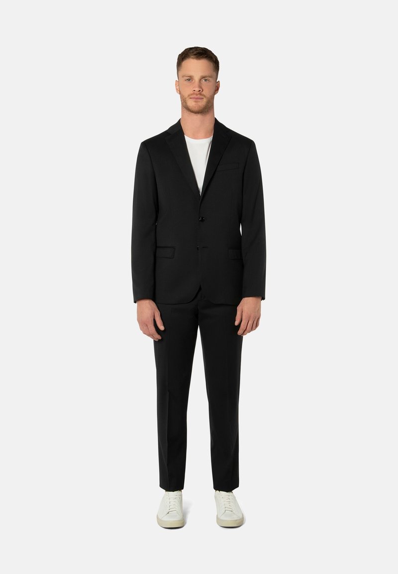 Schwarzer Anzug mit maßgeschneiderter Passform, einreihigem Blazer mit einem Knopf, aufgesetzten Revers und Seitentaschen, kombiniert mit weißem T-Shirt und lässigen weißen Sneakers.