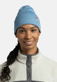 Gorro de punto azul claro con textura acanalada, que presenta un parche con un logo. Usado con una chaqueta de forro polar crema con acentos y botones grises.