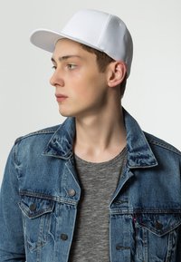 White cap with mesh back, curved brim, worn with a denim jacket and gray striped t-shirt. Close-up profile view showcasing the cap’s design.