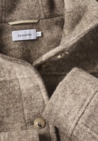 Giacca in feltro marrone chiaro con colletto, caratterizzata da due bottoni e una superficie texturizzata. Include un'etichetta di cura all'interno.