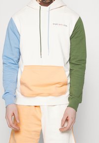 Väriblokki huppari valkoisena, sinisenä, vihreänä ja persikkana; siinä on edessä tasku, logon kirjailu ja kontrastiväriset nyörit.