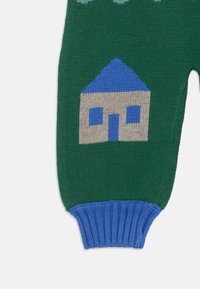 Pleteni zeleni kos, ki ima modre rebraste manšete; na sprednji strani je zasnova hiše v modri in bež barvi, z teksturirano, vzorčasto površino.