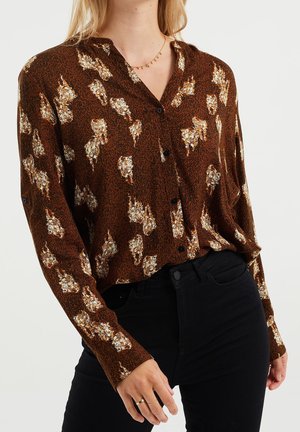 Vrouw die een bruin overhemd met lange mouwen draagt, met witte bloemvlekken en zwarte broek, afgebeeld van nek tot heupen.