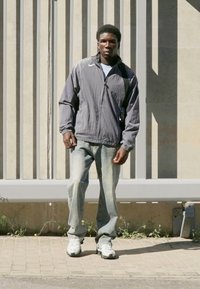 Nike vindjakke i mørkegrå, løstsiddende blå jeans og hvide sneakers. Har stående krave og lynlås lukning.