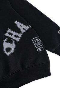 Sweatshirt noir avec un grand texte gris, un logo et une étiquette d'entretien. Col rond classique et poignets côtelés. Le matériau semble doux.
