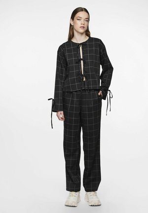 Chaqueta corta con patrón de cuadros negros y lazos en el frente, combinada con pantalones a juego con el mismo patrón. Se complementa con zapatillas deportivas voluminosas de color claro.
