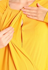 Gelbe Bluse mit Knopfleiste, mit drapiertem Stoff, der einen gefältelten Effekt am Ausschnitt erzeugt, und einer weichen, glatten Textur.
