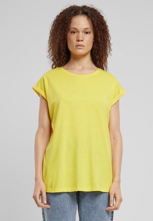Maglietta gialla a maniche corte con scollatura rotonda, vestibilità ampia e maniche arrotolate; abbinata a jeans in denim azzurro chiaro. Tessuto dalla consistenza liscia.