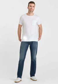 T-shirt en coton blanc avec une poche avant et jeans en denim bleu. Modèle portant des baskets blanches, se tenant droit devant un arrière-plan uni.