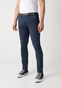 Tmavě modré úzké chinos kalhoty z bavlny s hladkou texturou. Kombinovány s černými plátěnými teniskami a černým pásem s vzorovaným povrchem.