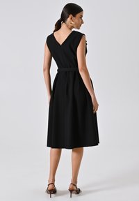Zwarte mouwloze jurk met een V-vormige rug, aansluitend lijfje, ceintuur in de taille en een vloeiende rok. Gemaakt van een gestructureerde stof met een gladde textuur.