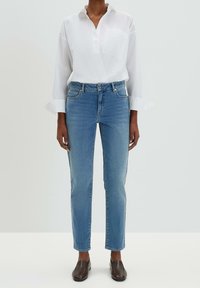 Valkoinen pitkähihainen paita, jossa on paikkatasku, yhdistettynä vaaleansinisiin suoralahkeisiin farkkuihin ja tummanruskeisiin slip-on-kenkiin.